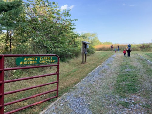 A gravel path leads into a nature sanctuary, with a sign for Audubon Sanctuary and people walking in the distance.