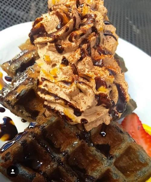 A stack of waffles topped with whipped cream, chocolate drizzle, and a slice of strawberry on the side.