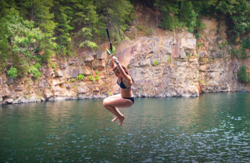 A person jumps off a rope swing into a lake surrounded by rocky cliffs and lush greenery.