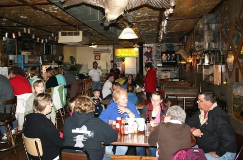 A busy restaurant interior with patrons at tables, a bar, and decor including a mounted bird overhead.