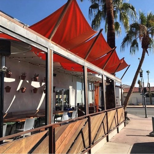 A modern restaurant patio with red awnings, wooden railings, and palm trees under a clear blue sky.