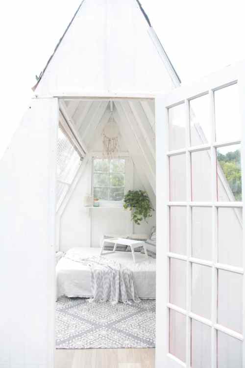 A bright, airy interior of a white A-frame cabin with a bed, plants, and large windows.