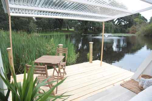 A wooden deck with a table and chairs overlooks a calm pond surrounded by greenery and a fountain in the background.