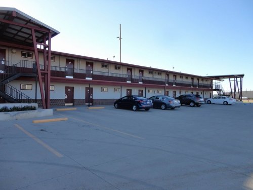 A low-rise motel with a parking lot, featuring multiple rooms and a clear blue sky in the background.