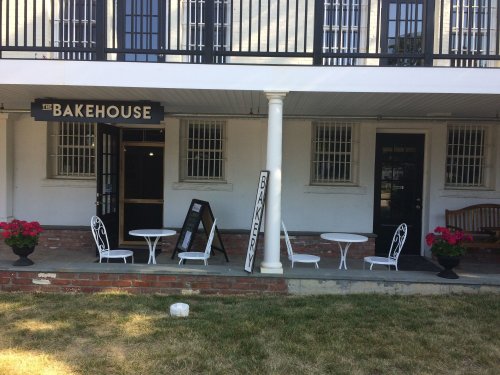 A quaint bakery entrance with white tables and chairs, potted flowers, and a sign reading "The Bakehouse."