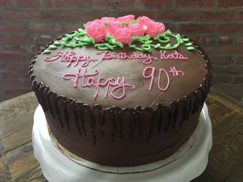 A chocolate birthday cake with pink roses and icing, celebrating a 90th birthday for "Kota."