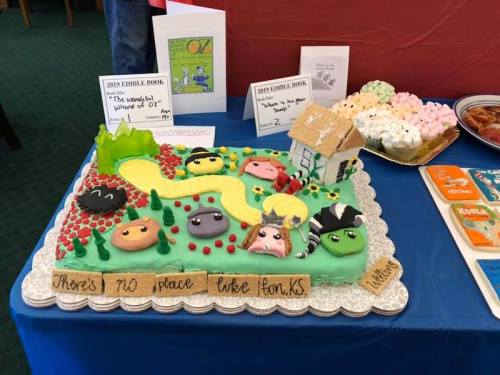 A colorful cake decorated with characters and a house, featuring a yellow brick road and the phrase "There's no place like home."