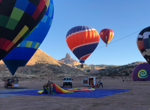 Colorful hot air balloons inflate against a clear sky, with people preparing a balloon on the ground in a scenic landscape.