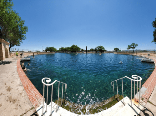 A clear blue swimming pool surrounded by trees and a sunny sky, with people swimming and relaxing.
