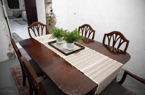 A wooden dining table set with a neutral runner and potted plants, surrounded by matching chairs in a minimalist room.