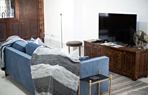 Cozy living room with a blue sofa, a wooden TV stand, and a fan, featuring a textured throw blanket and decorative pillows.