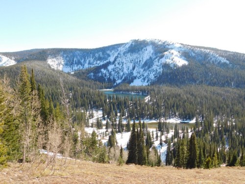 A scenic view of a mountainous landscape with a lake surrounded by evergreen trees and patches of snow.