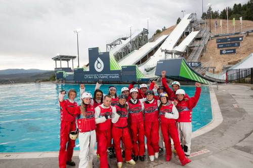 A group of athletes in red suits celebrates at Utah Olympic Park, with ski jumps and a pool in the background.