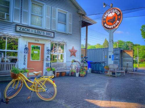 A yellow bicycle with a plant sits outside Zach & Lou's BBQ, featuring a sign and a charming, rustic building.
