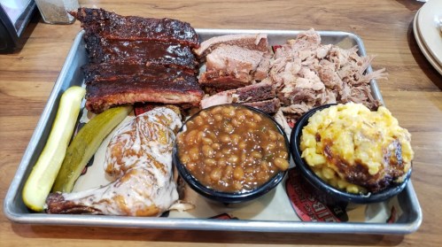 A metal tray with barbecue ribs, pulled pork, chicken, baked beans, macaroni and cheese, and pickles.