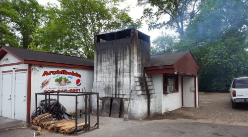 A brick building with a tall, blackened chimney, surrounded by trees and a parked vehicle. Smoke is rising from the chimney.