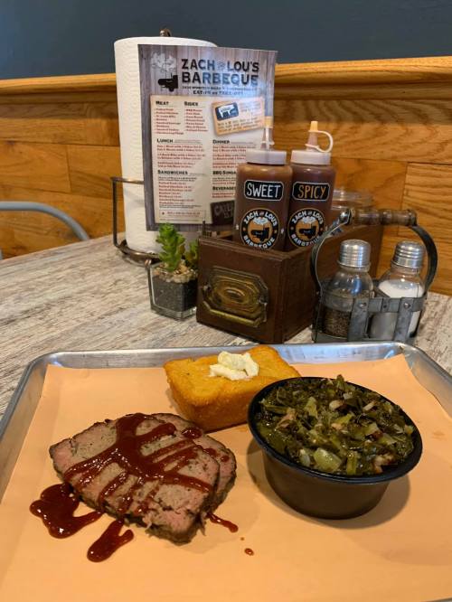 A tray with sliced barbecue brisket drizzled in sauce, cornbread, collard greens, and condiments in the background.