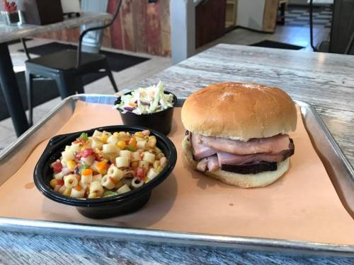 A tray with a sandwich featuring ham and a bun, alongside pasta salad and coleslaw in black bowls.