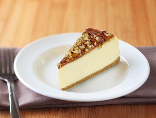A slice of cheesecake topped with nuts and caramel on a white plate, resting on a wooden surface.