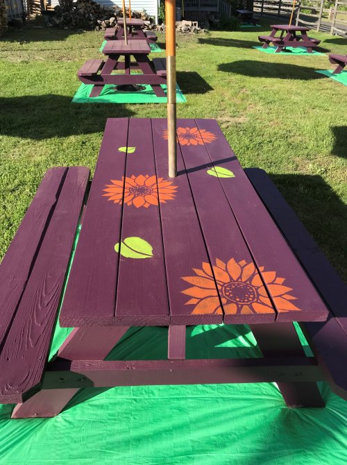 A purple picnic table with orange sunflower designs, set on green grass with other tables in the background.