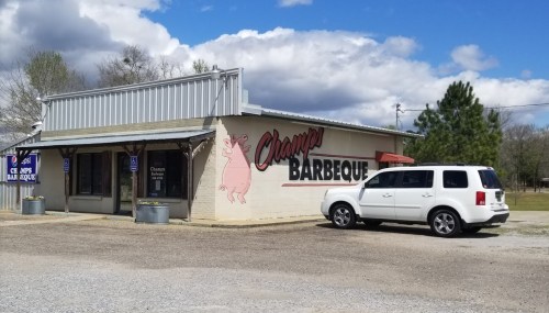 A barbecue restaurant with a pig mural on the wall, featuring a white SUV parked outside under a cloudy sky.