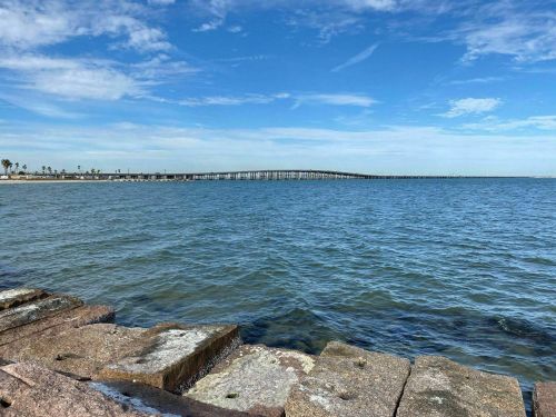 A calm sea view with a pier in the distance under a blue sky, featuring rocky shoreline in the foreground.