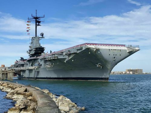 A large aircraft carrier docked near a rocky shoreline under a clear blue sky.