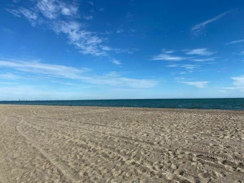 A sandy beach stretches towards a calm blue sea under a clear sky with scattered clouds.