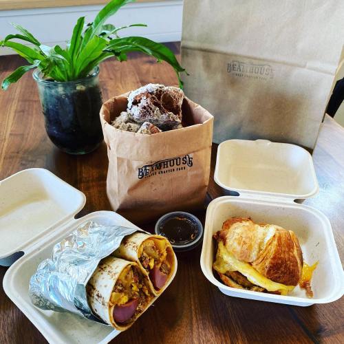 A table with takeout food: a wrapped burrito, a croissant sandwich, a bag of pastries, and a plant in a pot.