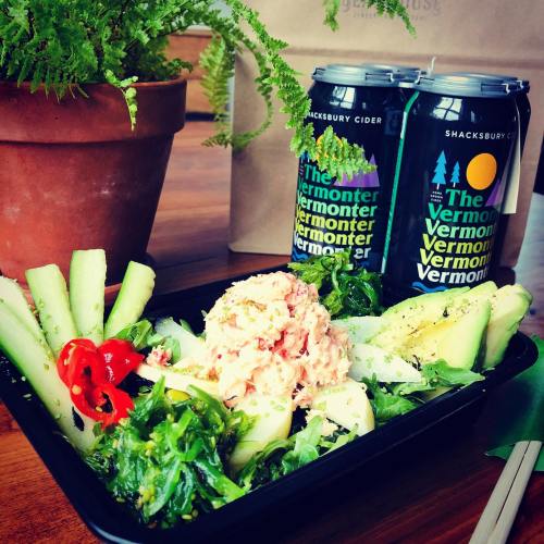 A fresh salad with greens, cucumber, and a scoop of seafood salad, alongside two cans of Shacksbury cider.