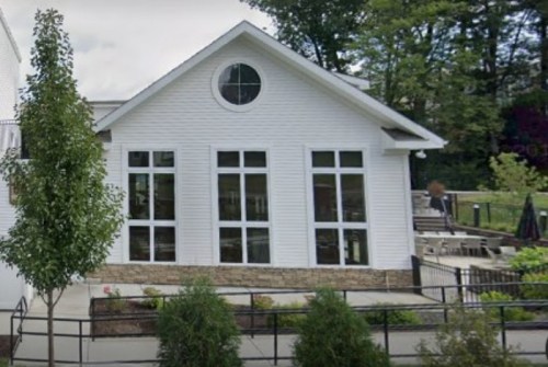 A modern white building with large windows, surrounded by greenery and a patio area.