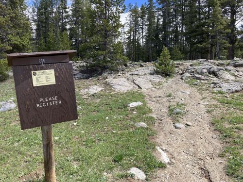 A wooden registration box on a trail surrounded by rocks and trees, with a clear sky above.