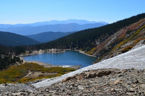 A serene mountain landscape featuring a blue lake surrounded by trees and distant mountains under a clear blue sky.