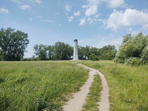 A grassy path leads to a tall lighthouse surrounded by trees under a blue sky with scattered clouds.