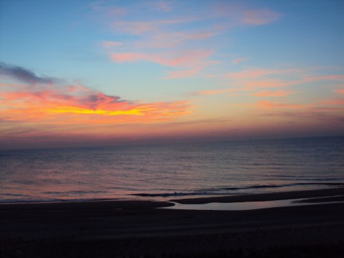 A serene beach at sunset, with vibrant orange and pink hues reflecting on the calm ocean waters.