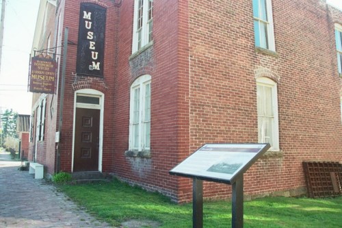 A brick building with a sign reading "Museum" and an informational plaque in front on a sunny day.