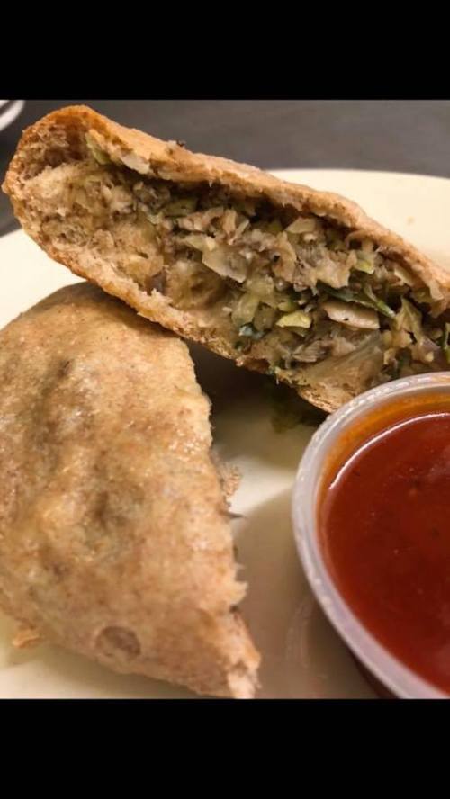 A close-up of a stuffed pastry with a filling of vegetables, served with a small cup of red sauce.