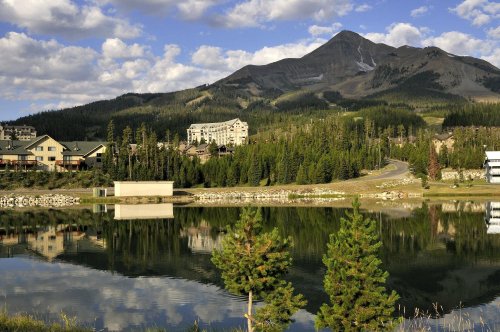 A serene mountain landscape with a lake reflecting trees and buildings under a blue sky with fluffy clouds.