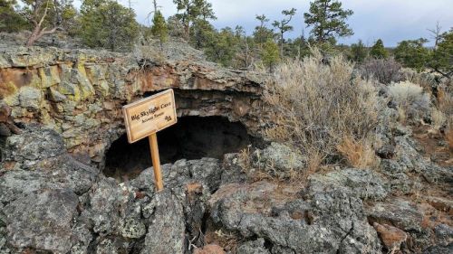A rocky cave entrance with a wooden sign reading "Big Spring Cave" surrounded by sparse vegetation and trees.