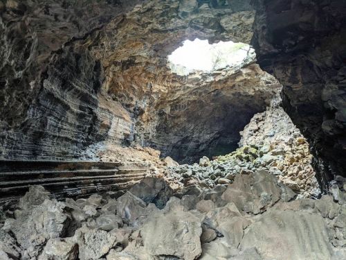 A rocky cave interior with a large opening at the top, allowing light to filter in and illuminate the stone formations.