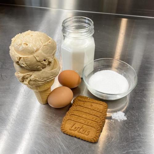 A cone of ice cream, a jar of milk, two eggs, a bowl of sugar, and Lotus cookies on a stainless steel surface.