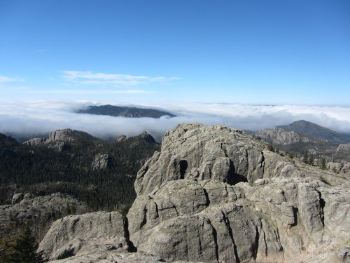 A rocky mountain peak overlooks a sea of clouds and distant hills under a clear blue sky.