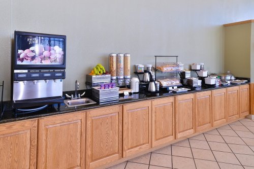 A breakfast buffet featuring a juice dispenser, cereal bins, pastries, and fruit on a wooden counter.