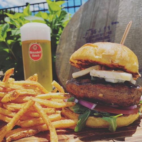 A juicy burger with toppings and fries, accompanied by a cold beer, set against a sunny outdoor backdrop.