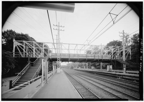 Black and white image of a railway bridge with stairs, surrounded by trees and power lines, viewed from a train platform.