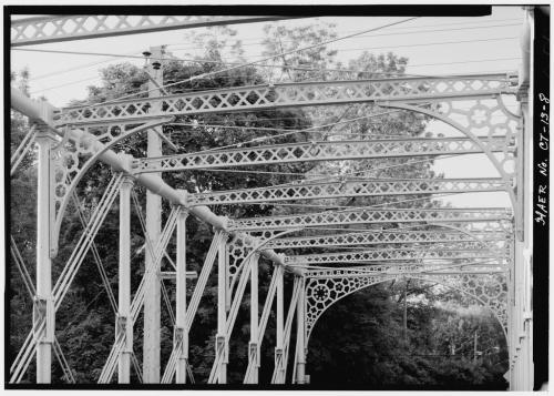 Black and white image of a metal bridge's intricate latticework and arches, surrounded by trees.