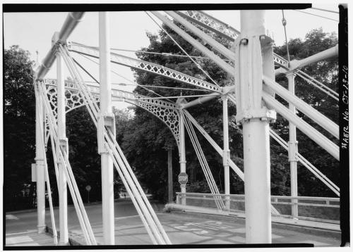 Black and white image of a decorative metal bridge structure with trees in the background and a stop sign visible.