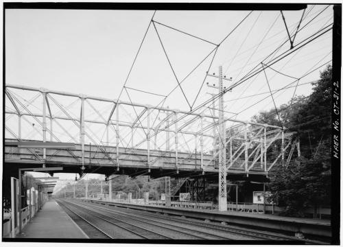 Black and white image of a railway bridge with overhead wires, viewed from a train platform.