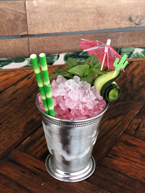 A silver cup filled with crushed ice, pink drink, mint leaves, a green straw, and decorative garnishes.
