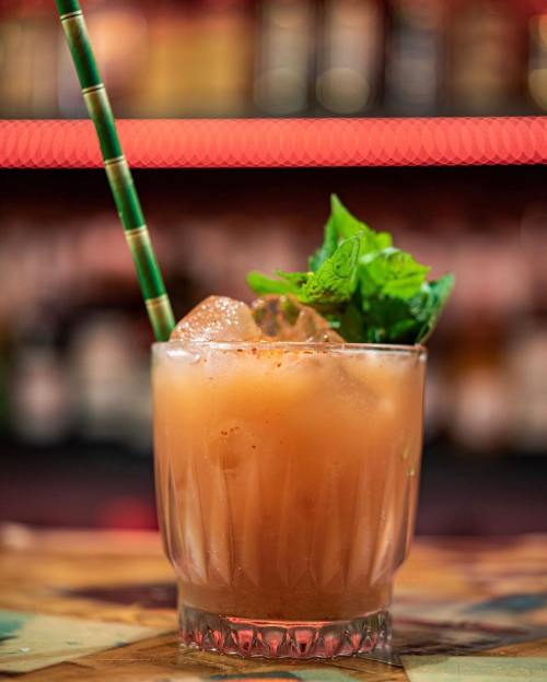 A refreshing cocktail in a glass with ice, garnished with mint leaves and a green straw, set against a blurred bar background.